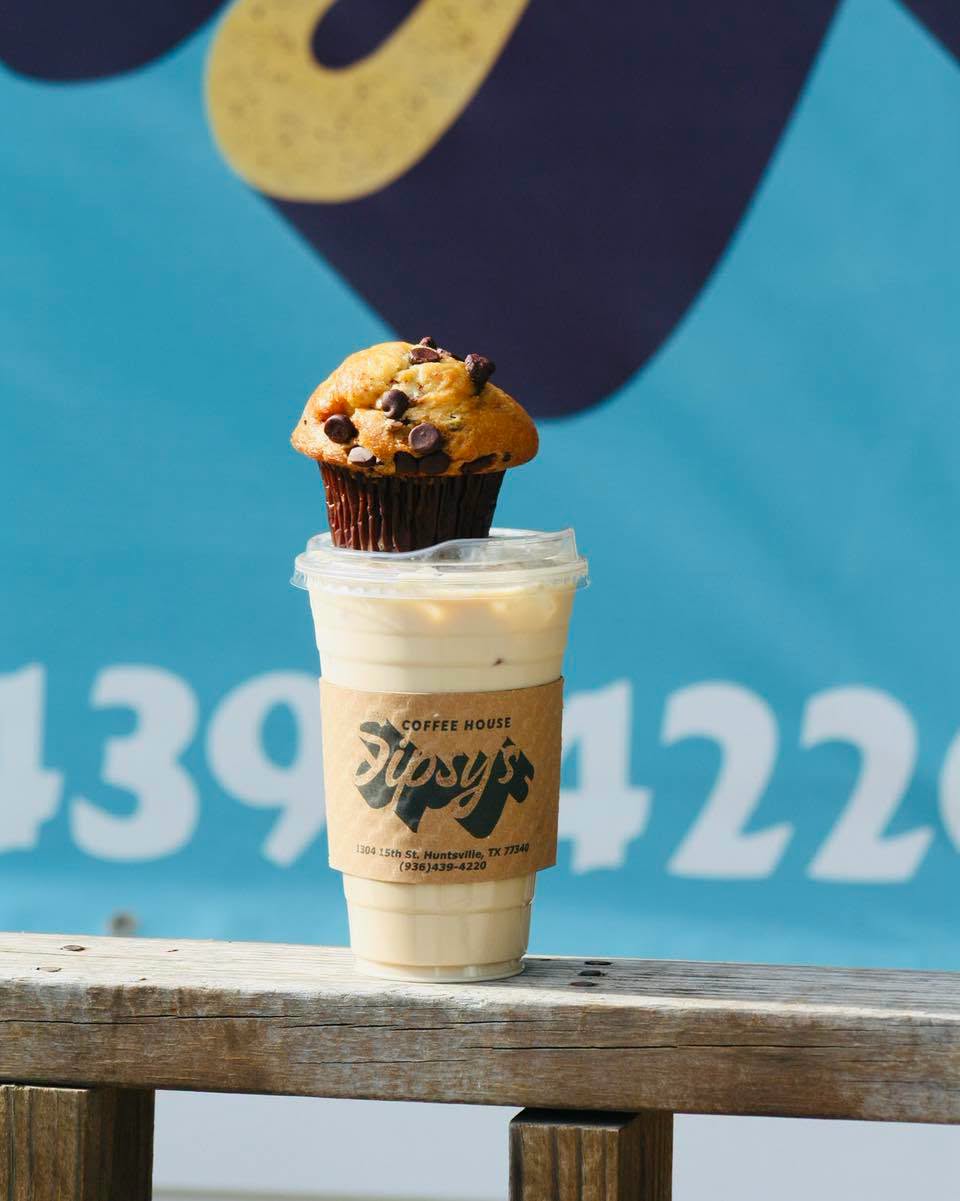 Sipsy's iced latte in a branded cup with a chocolate chip muffin resting on the lid, on a wooden railing in front of a blue food truck